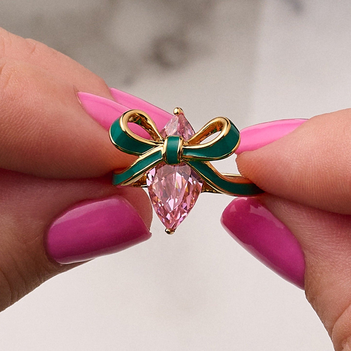 Close-up of Amelia Bow Ring with blush pink cubic zirconia and hand-painted emerald green enamel bow on recycled gold plating, held by hands with pink nails