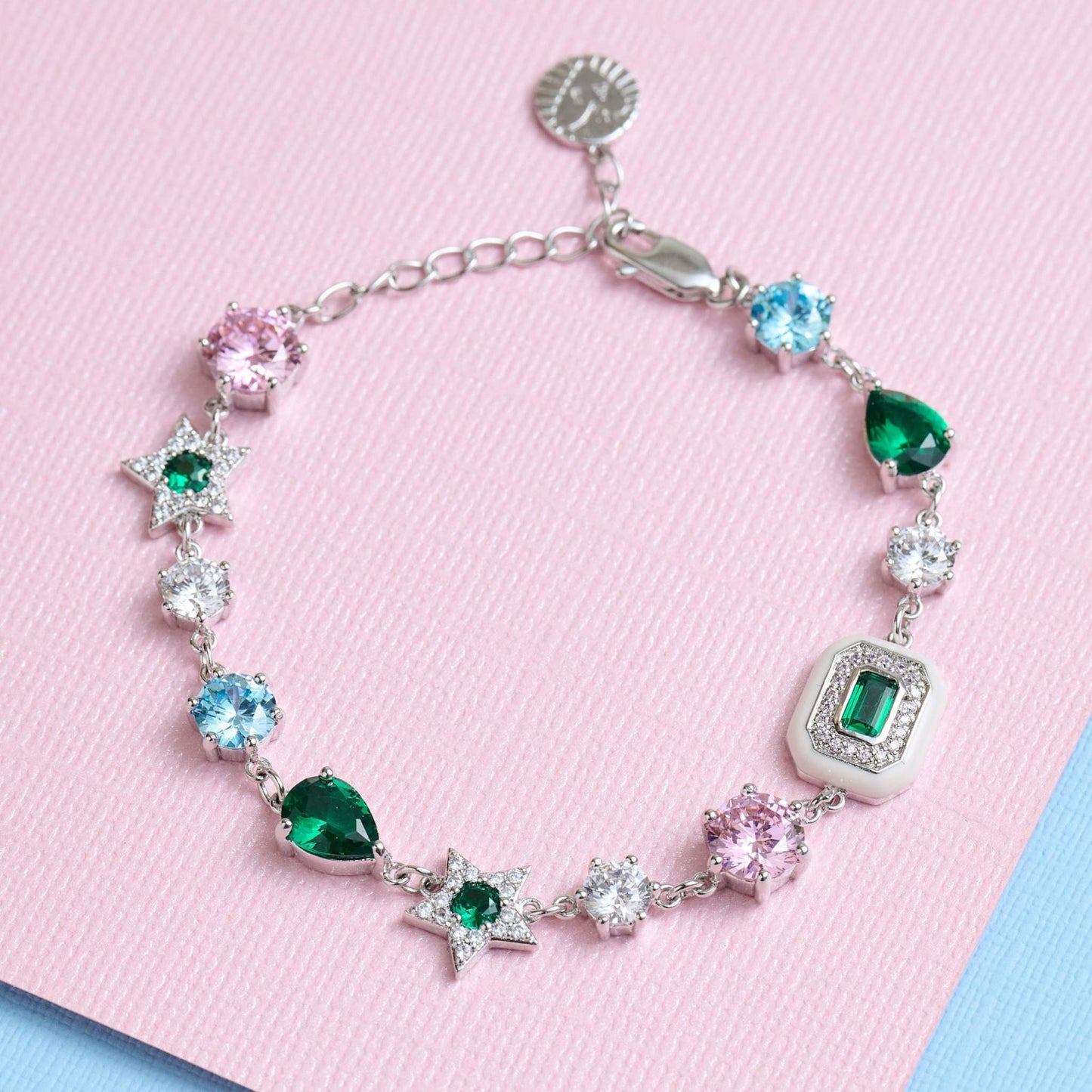 Celeste Cluster Bracelet with blush pink enamel charms, emerald green cubic zirconia, and silver recycled brass chain on pink background