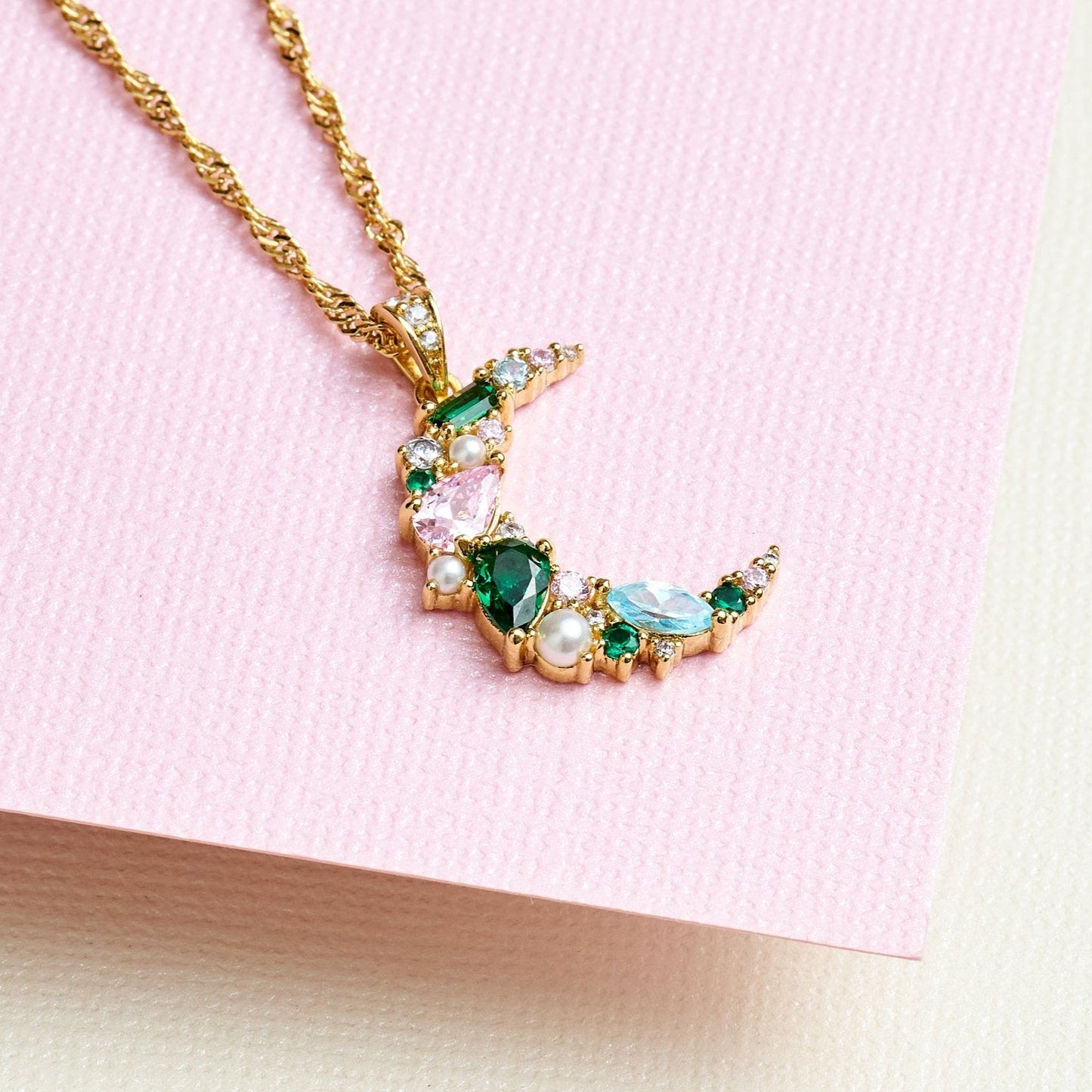 Celeste Cluster Moon Pendant necklace with blush pink, emerald green stones, faux pearls, and gold chain on pink background