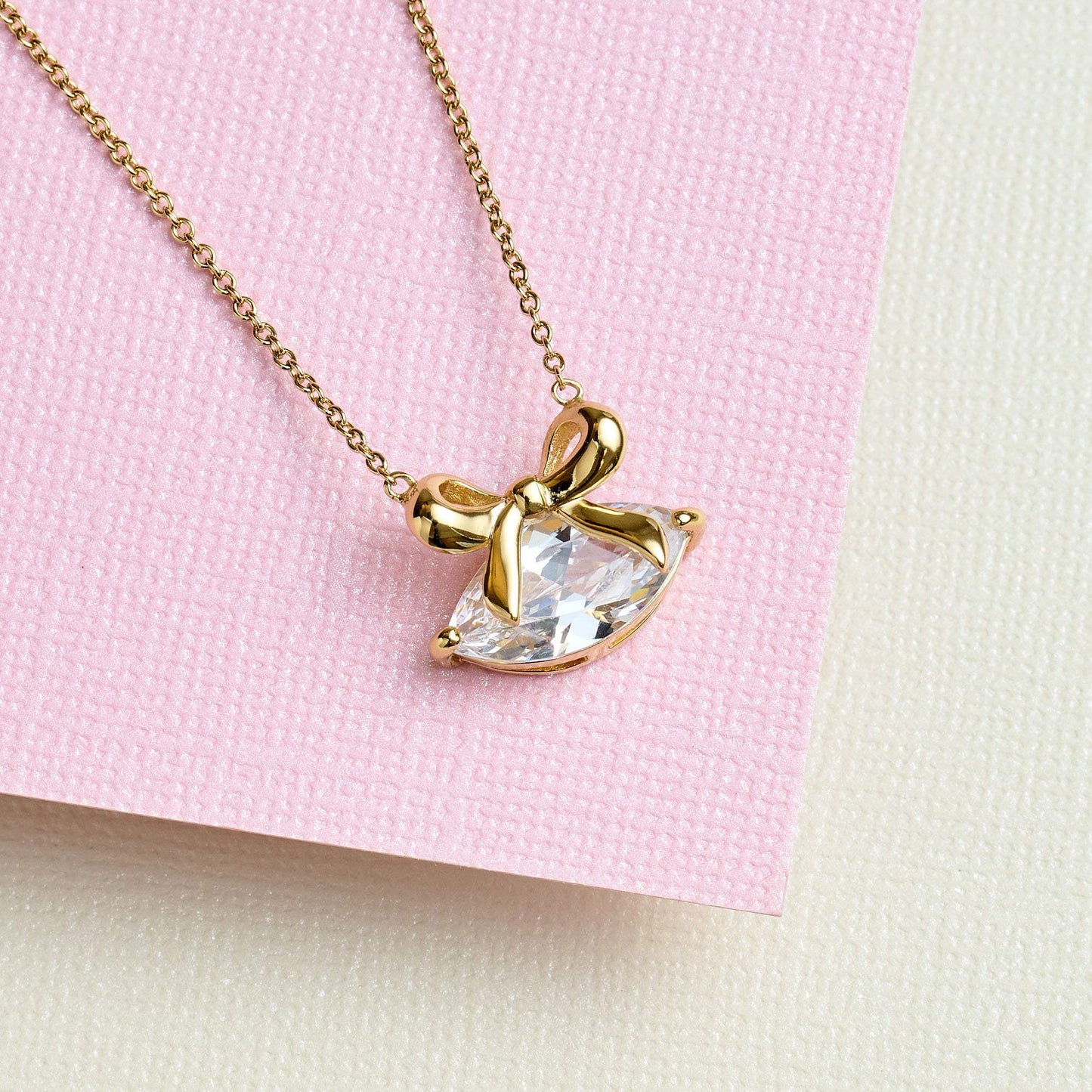 Amelia Bow Necklace gold chain with clear marquise cubic zirconia bow pendant on pink textured background