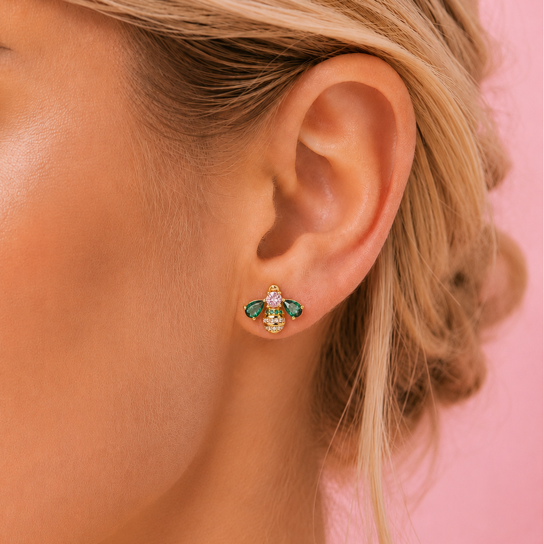 A close-up of a woman's ear wearing a small gold bumble bee stud earring with green and clear rhinestones, set against a solid pink background.