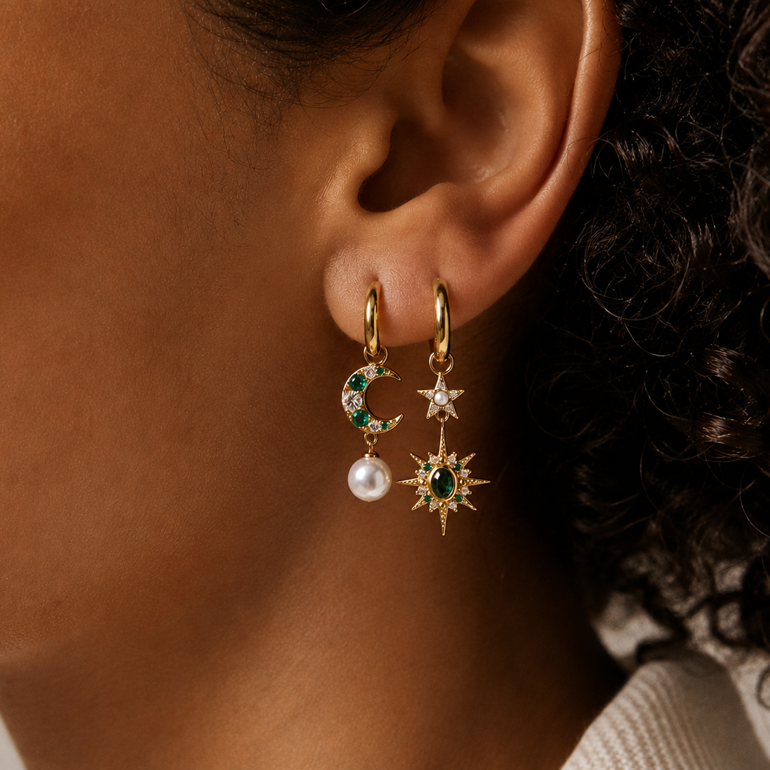 Close-up of celestial moon and star gold huggie hoop earrings with emerald green gemstones and pearls.
