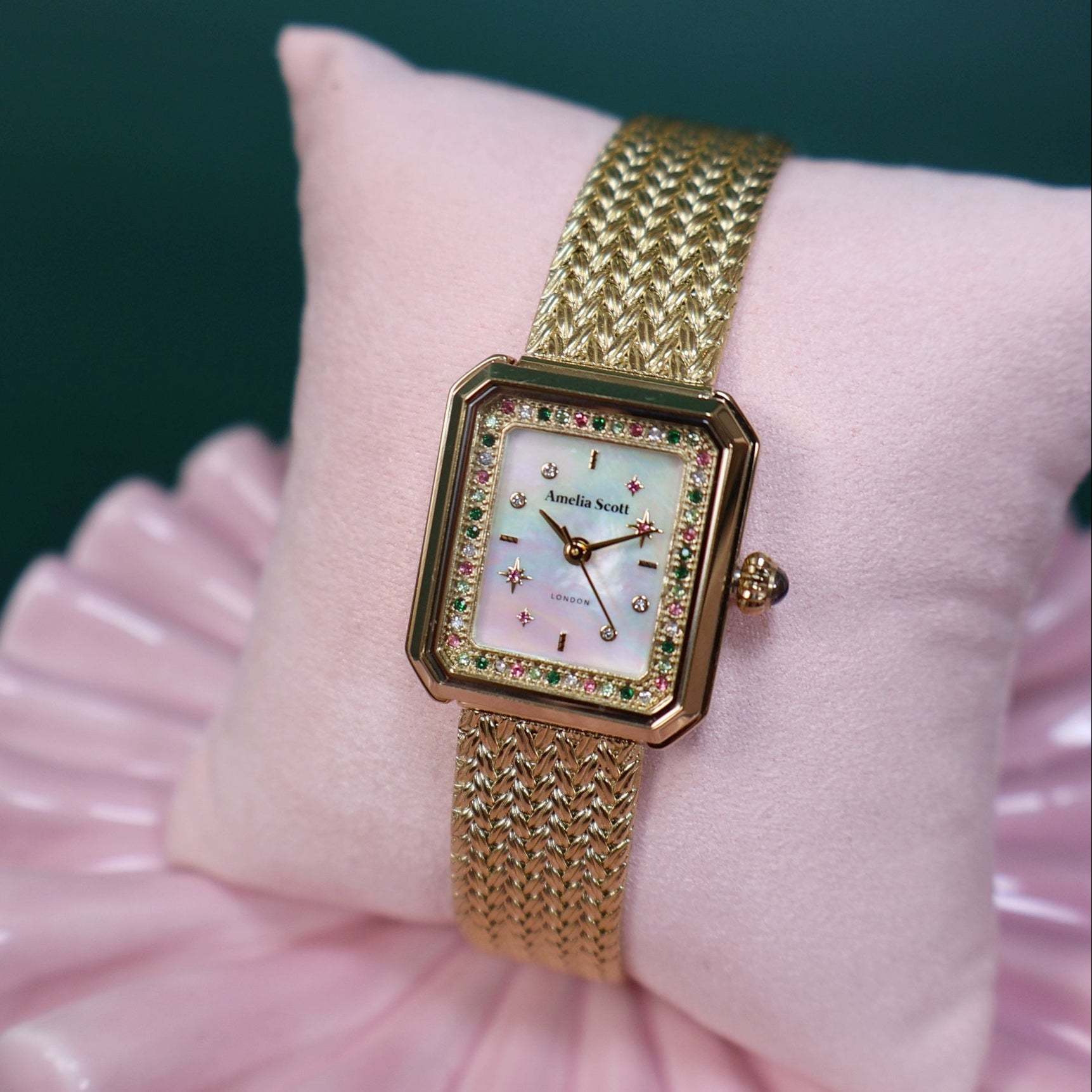 Gold vintage style ladies watch with a gold mesh strap on a pink decorative shell against a dark emerald background