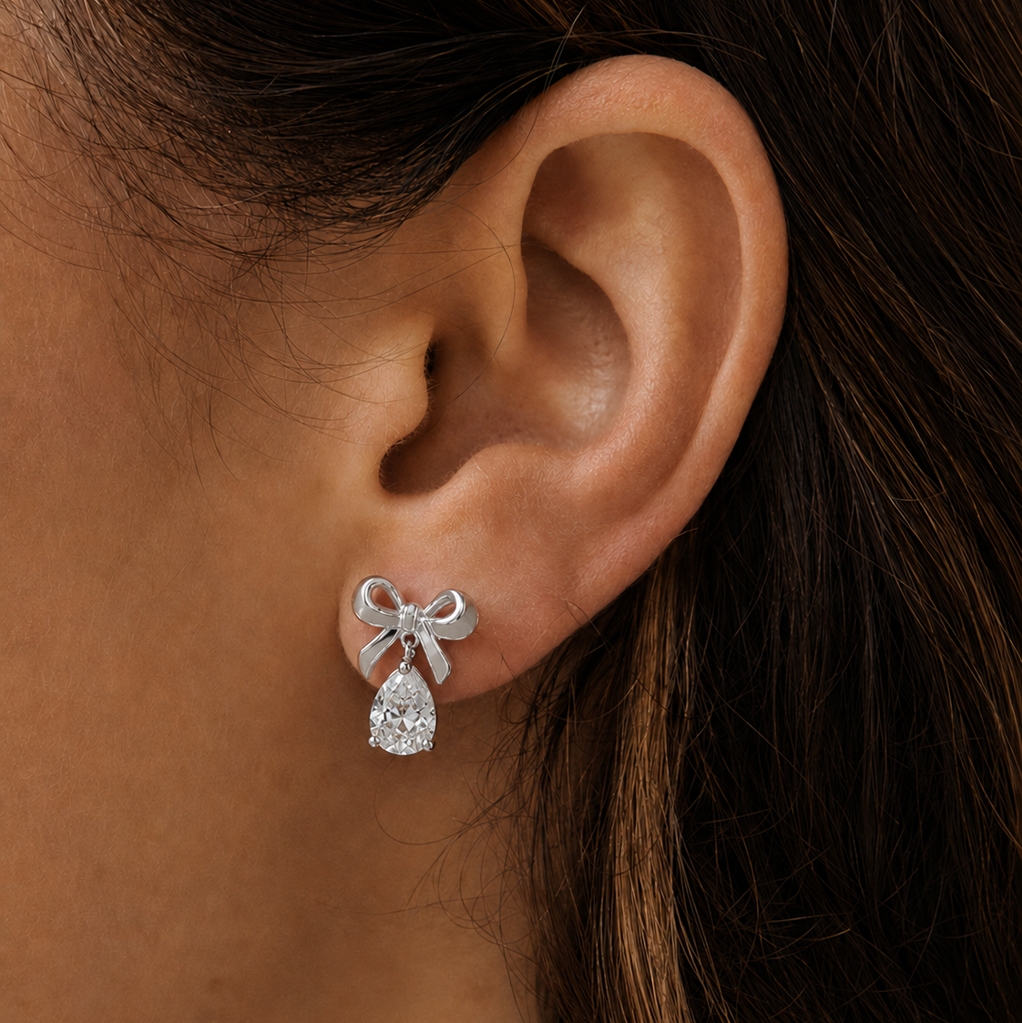 A close-up of a model's ear wearing a silver bow stud earring with a clear teardrop-shaped crystal pendant.