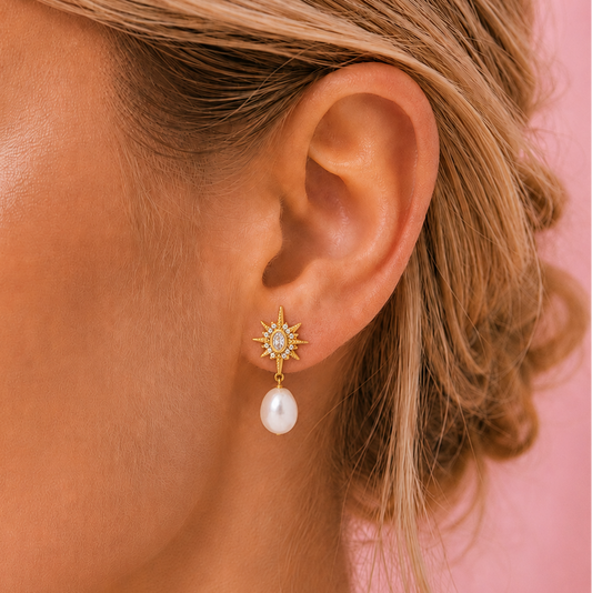 A close-up of a woman's ear wearing a gold starburst stud bridal pearl earring with a dangling white pearl, set against a soft pink background.