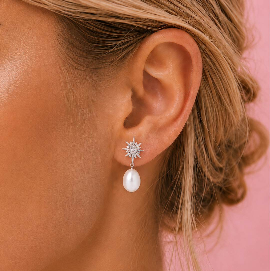 A close-up of a woman's ear wearing a dangling earring with a silver sunburst stud and a white teardrop pearl, set against a solid pink background.