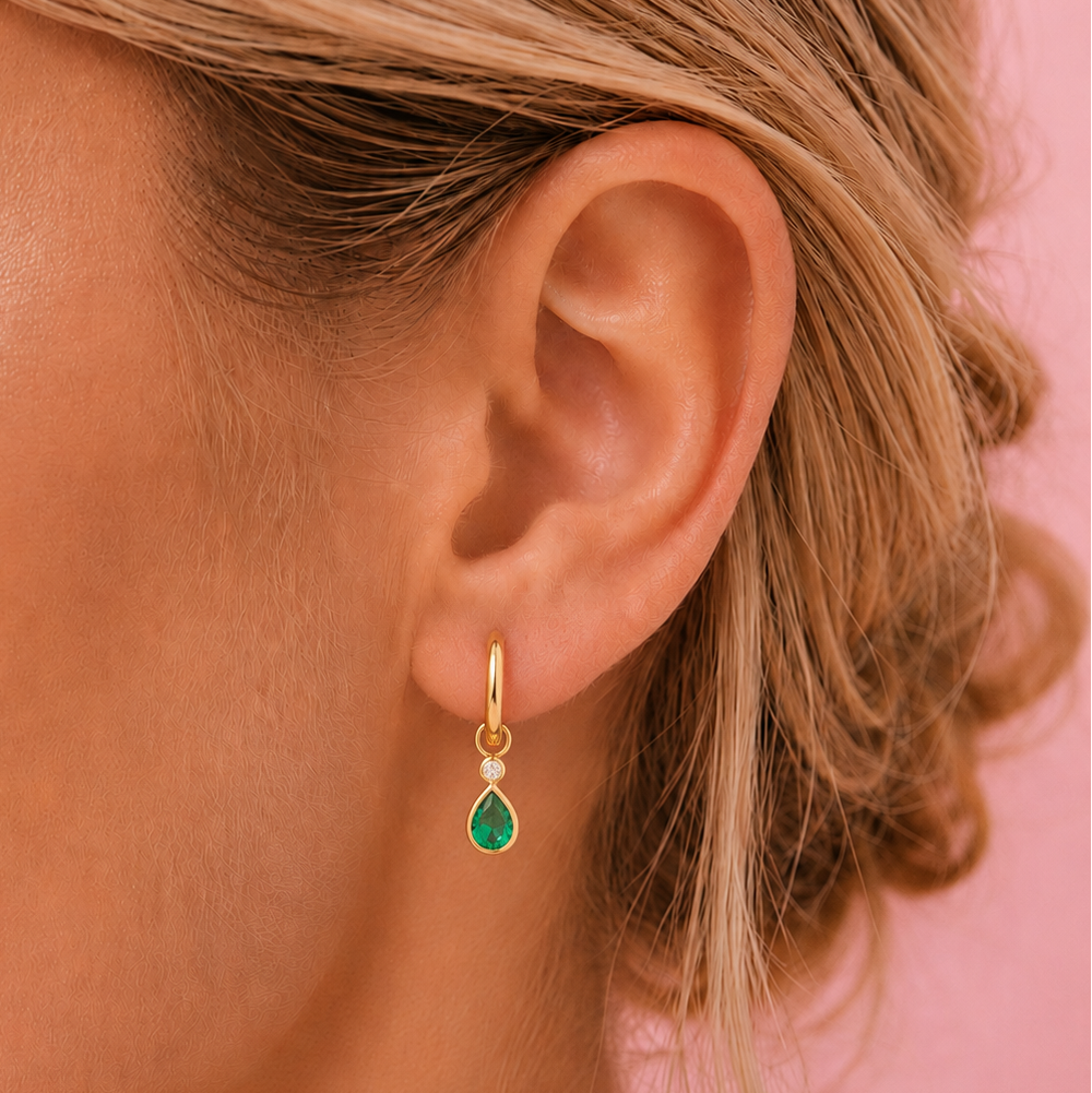 A close-up of a model's ear wearing a small gold huggie hoop earring with a teardrop-shaped green gemstone, set against a solid pink background.