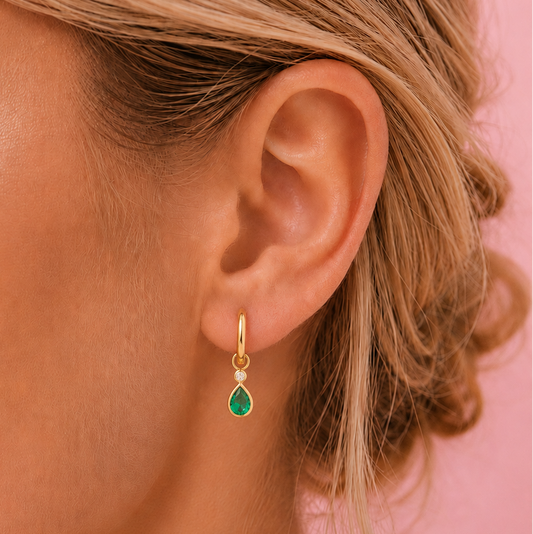 A close-up of a model's ear wearing a small gold huggie hoop earring with a teardrop-shaped green gemstone, set against a solid pink background.