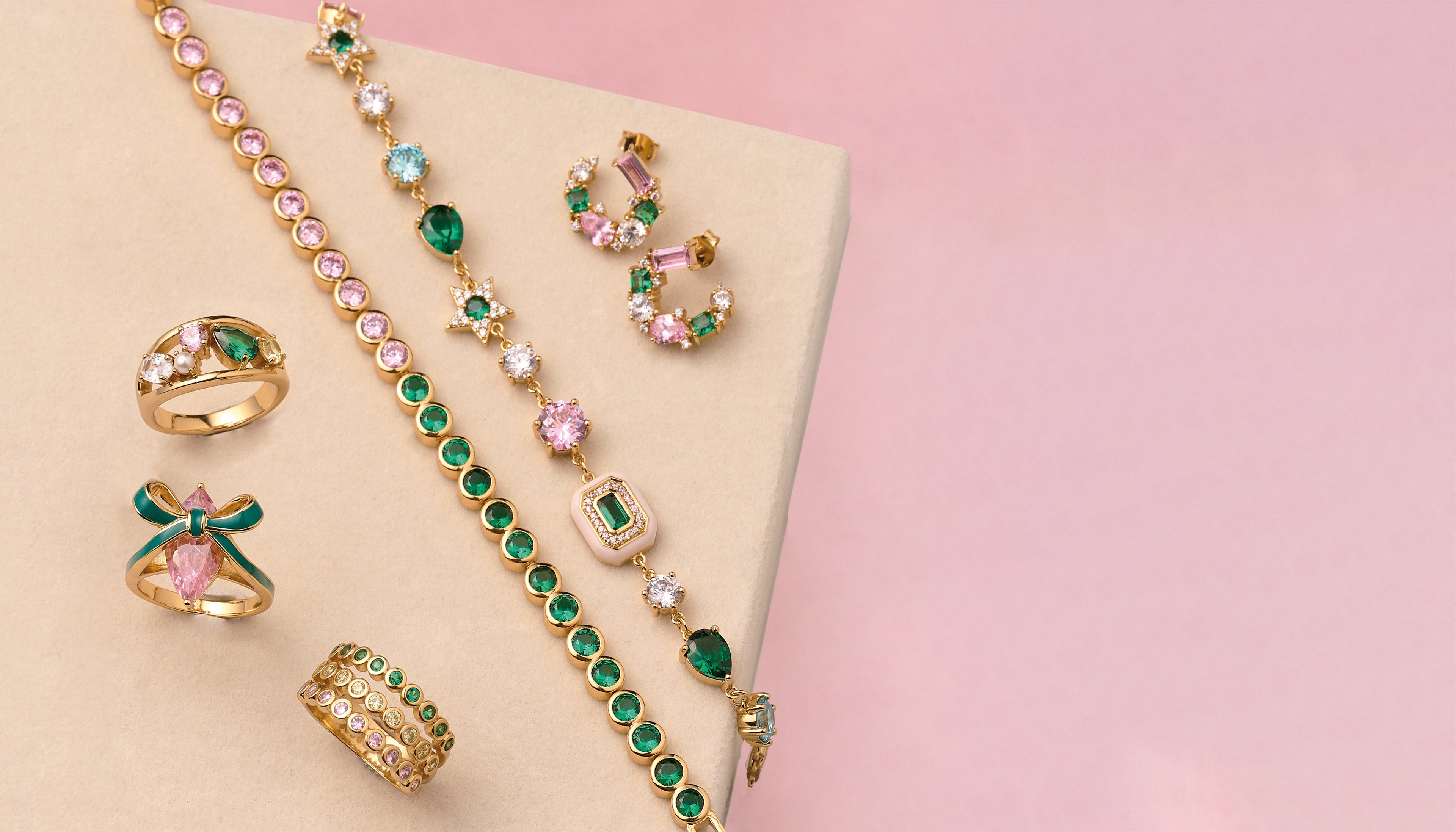Gold jewelry with green and pink gemstones on a beige and pink background