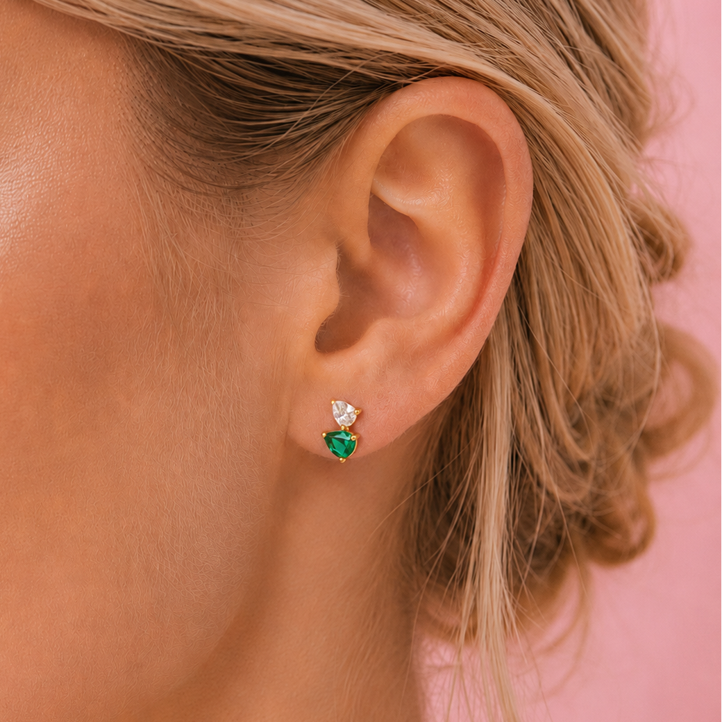 A close-up of a model's ear wearing a small dangle earring with a green heart-shaped gemstone and a small white heart-shaped gemstone above it, set against a solid pink background.